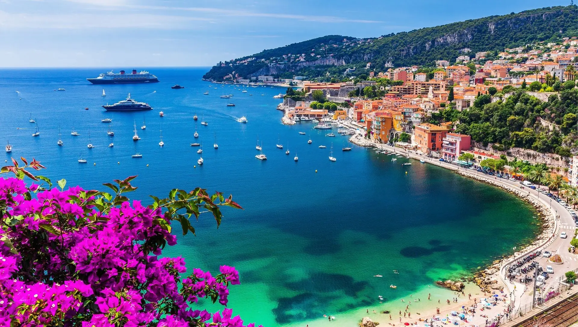 Vue panoramique de la baie de Villefranche-sur-Mer sur la Côte d'Azur, avec des yachts et bateaux à voile ancrés dans une mer turquoise, des bâtiments colorés en bord de colline, des plages fréquentées et des bougainvilliers en fleurs au premier plan.